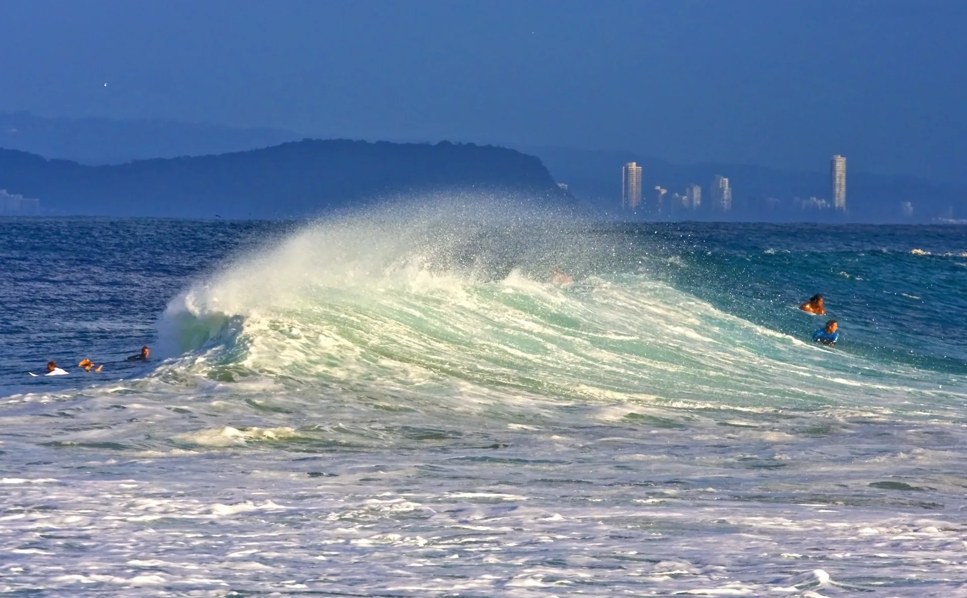 A wave curling over and breaking at Rainbow Bay, Coolangatta, with a headland and Gold Coast high-rises behind